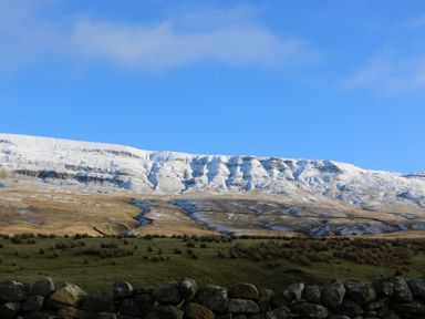 A landscape with a stone wall in the foreground green grass patches and a snow-covered hill under a blue sky at The Folly in Linton near Thresfield
