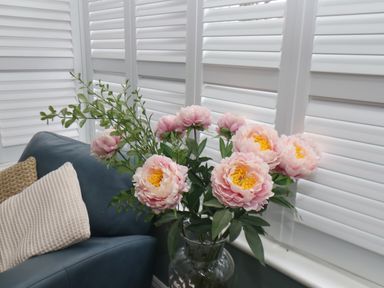 A bouquet of pink flowers in a glass vase next to a blue chair with two cushions and white shutters in the background at South Bay Beach House in Bridlington