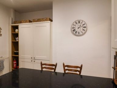 A kitchen area with a black countertop table, two wooden chairs, a white cabinet with shelves holding kitchen items, a fire extinguisher, and a wall clock at South Bay Beach House in Bridlington