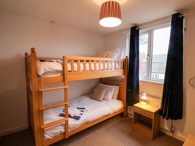 A bedroom with a wooden bunk bed next to a window with dark curtains and a wooden nightstand with a lamp at South Bay Beach House Bridlington