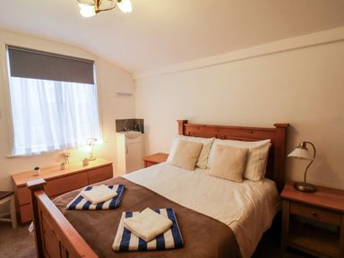 A bedroom with a wooden bed with pillows and towels a bedside table with a lamp a dresser with plants and a window with a blind at South Bay Beach House in Bridlington