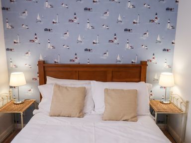 A bedroom with a wooden headboard bed two beige pillows white sheets two wicker side tables with lamps and wallpaper with lighthouses and sailboats at South Bay Beach House in Bridlington