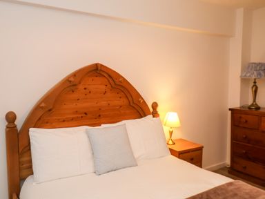 A bedroom with a wooden bed frame white pillows a bedside table with a lamp and a wooden dresser with a lamp at South Bay Beach House in Bridlington