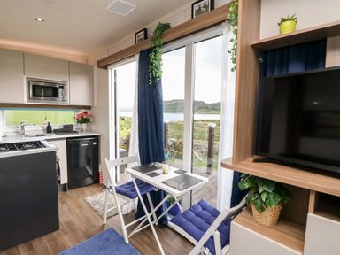 A small dining table with two chairs and blue cushions next to sliding glass doors in a kitchen with appliances and a television on a wooden stand with plants at Balvicar Beag in Balvicar Isle Of Seil