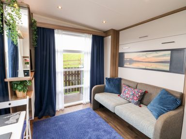 A living room with a sofa cushions a blue rug a window with curtains and a desk with a plant at Balvicar Beag in Balvicar Isle Of Seil