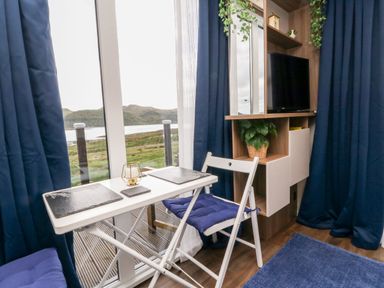 A dining area with a white table and two chairs next to a window with blue curtains and a shelving unit with a TV and plants at Balvicar Beag in Balvicar Isle Of Seil