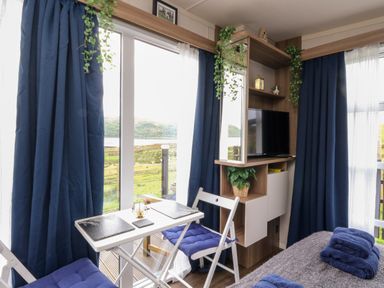 A small dining area with two chairs and table near large windows with blue curtains and a corner unit with a television and plants at Balvicar Beag in Balvicar Isle Of Seil