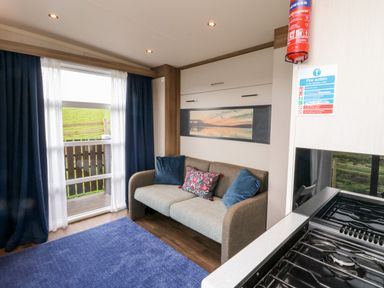 A small living area with a sofa with cushions a blue rug a window with curtains and a kitchen stove at Balvicar Beag in Balvicar Isle Of Seil