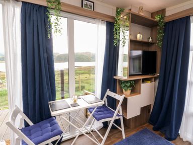 A small dining area with two chairs and a table by sliding glass doors next to a corner shelf with a television and plants at Balvicar Beag in Balvicar Isle Of Seil