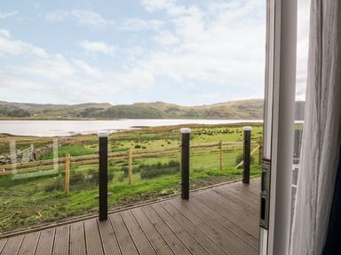 A view of a grassy field and hills across water seen from a wooden deck with glass railings at Balvicar Beag in Balvicar Isle Of Seil