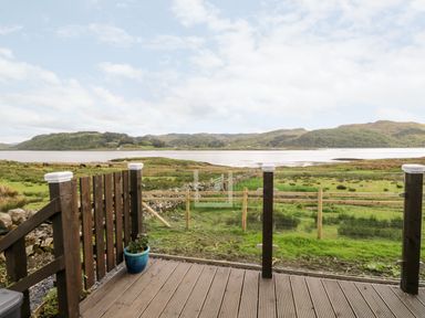 A wooden deck with a potted plant overlooking a grassy area and a body of water with hills in the background at Balvicar Beag Balvicar Isle Of Seil