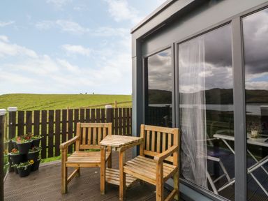A patio with two wooden chairs and a small table near sliding glass doors overlooking a grassy hill at Balvicar Beag in Balvicar Isle Of Seil