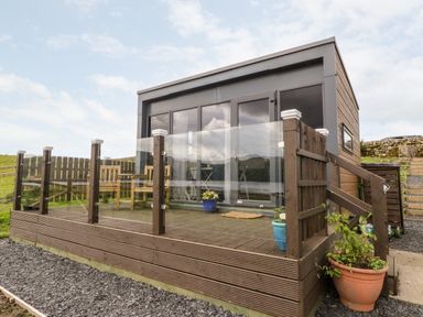 A wooden deck with glass railing and potted plants outside a small modern cabin at Balvicar Beag in Balvicar Isle Of Seil