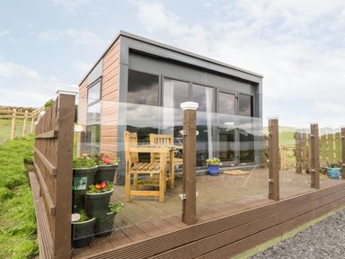 A wooden deck with glass railing and wooden chairs outside a modern building at Balvicar Beag in Balvicar Isle Of Seil