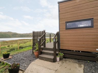 A wooden house with steps and potted plants outside next to a fenced grassy area and a body of water at Balvicar Beag Balvicar Isle Of Seil
