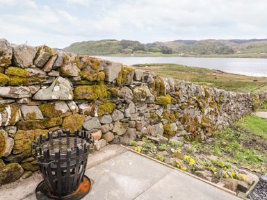 A stone wall with moss beside a patio and a metal fire basket with a view of hills and water at Balvicar Beag in Balvicar Isle Of Seil