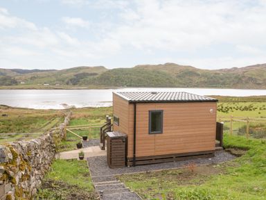 A small wooden cabin near a body of water with hills in the background at Balvicar Beag Balvicar Isle Of Seil