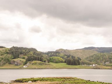 A lake with hills and trees in the background and a small boat on grassy land at Balvicar Beag in Balvicar Isle Of Seil