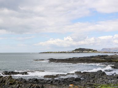 A rocky coastal shoreline with waves and a distant view of an island with buildings at Balvicar Beag Balvicar Isle Of Seil