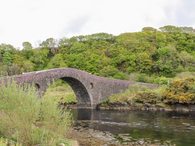 An old stone arch bridge over a river with green trees on a hillside at Balvicar Beag in Balvicar Isle Of Seil