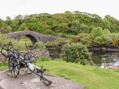 A stone arch bridge over a river with green trees and bushes around and an old black metal agricultural machine on a concrete area near green grass at Balvicar Beag in Balvicar Isle Of Seil