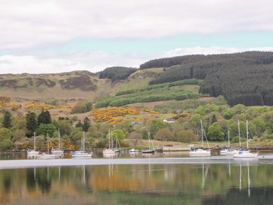 A body of water with sailboats anchored near a shore with trees and hills in the background at Balvicar Beag Balvicar Isle Of Seil