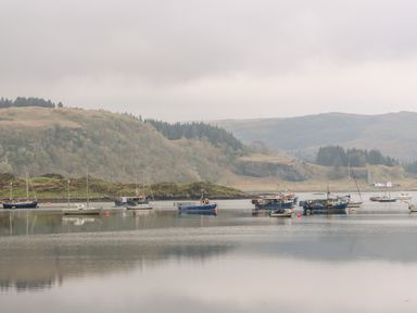 Boats moored on calm water with hills and trees in the background at Balvicar Beag Balvicar Isle Of Seil