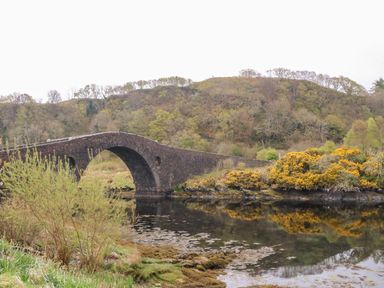 A stone arch bridge over a body of water with a wooded hill and bushes in the background at Balvicar Beag in Balvicar Isle Of Seil