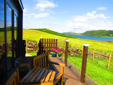 A wooden bench on a deck overlooking a grassy field and a body of water at Balvicar Beag in Balvicar Isle Of Seil