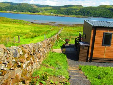 A small wooden cabin next to a stone wall with steps leading down to green fields and a body of water at Balvicar Beag Balvicar Isle Of Seil