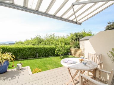 A patio with wooden table and chairs a watering can and plants overlooking a garden with trees and hedges at Blue Waters in Weymouth