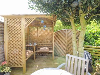 An outdoor wooden gazebo with a table cups and cushions beside a tree with string lights in a garden at Blue Waters in Weymouth