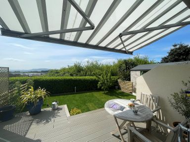 A patio with a round wooden table and two chairs under a striped awning overlooking a garden with green grass and bushes at Blue Waters in Weymouth