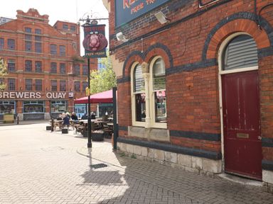 Outdoor seating area with people sitting at wooden tables outside a red brick building with a red door and a pub sign showing a lion near Brewers Quay building