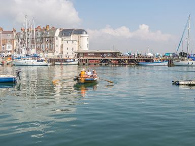 A small rowboat with people on a harbor with sailboats and buildings in the background at The Nightingale in Weymouth