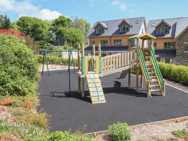 An outdoor playground with a slide swings and climbing ramp in front of residential buildings at 5 Court Cottage Hillfield Village in Dartmouth