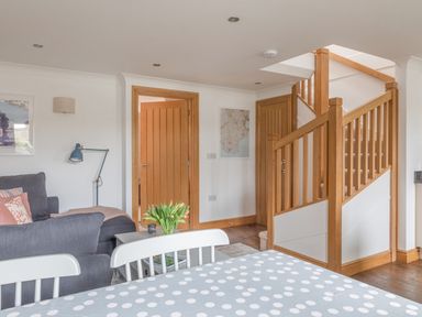 A room with a dining table covered by a polka dot tablecloth white chairs a staircase with wooden railings a grey sofa with cushions a floor lamp and wooden doors at 5 Court Cottage Hillfield Village Dartmouth