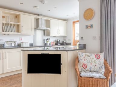 A kitchen with white cabinets a counter with utensils and appliances and a wicker chair with cushions at 5 Court Cottage Hillfield Village Dartmouth