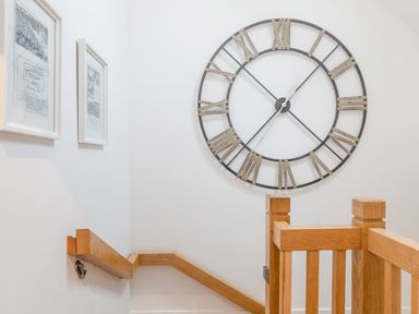 A staircase with wooden handrails a large wall clock with roman numerals and framed pictures on a white wall at 5 Court Cottage Hillfield Village in Dartmouth