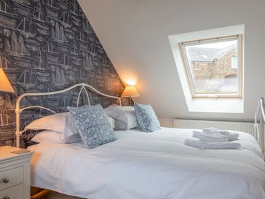 A bedroom with a neatly made bed with white linens and blue patterned pillows a nightstand with a lamp a window in the sloped ceiling and wallpaper with sailboat designs at 5 Court Cottage Hillfield Village Dartmouth
