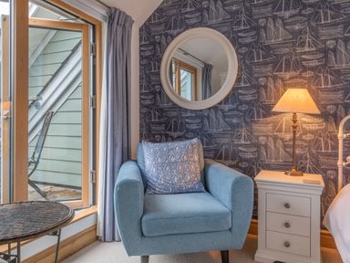 A room corner with a blue armchair and patterned cushion beside a white bedside table with a lamp and round mirror on a wall with boat designs at 5 Court Cottage Hillfield Village Dartmouth