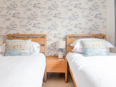 A bedroom with two single beds with white bedding a wooden nightstand with a lamp and boat-themed wallpaper at 5 Court Cottage Hillfield Village Dartmouth