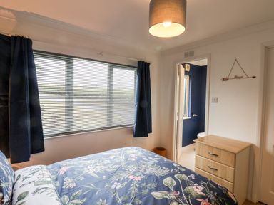 A bedroom with a bed, floral bedding, a window with blinds and dark curtains, a wooden chest of drawers, and a doorway leading to a bathroom at Handale Banks Farm Lodge in Liverton