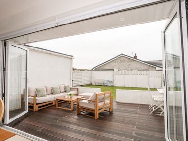 An outdoor patio area with wooden seating a wooden floor and a lawn with white fences at 31 Sisial Y Mor in Rhosneigr