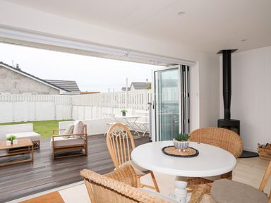 A dining area with a round white table and wicker chairs next to a black wood stove and an open patio with outdoor seating at 31 Sisial Y Mor in Rhosneigr