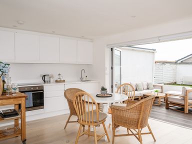 A kitchen and dining area with wicker and wooden chairs and a table opening to an outdoor seating area at 31 Sisial Y Mor in Rhosneigr