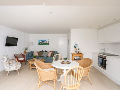 An open living room and kitchen with a round dining table surrounded by wicker and wooden chairs a blue sofa two armchairs a wall-mounted television and white kitchen cabinets at 31 Sisial Y Mor in Rhosneigr
