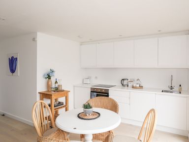 A kitchen with white cabinets a round dining table with four chairs and a wooden side table with flowers at 31 Sisial Y Mor in Rhosneigr