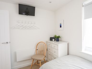 A small bedroom with a bed a wooden chair a white chest of drawers with a plant and radio hangers on the wall and a wall-mounted television at 31 Sisial Y Mor in Rhosneigr