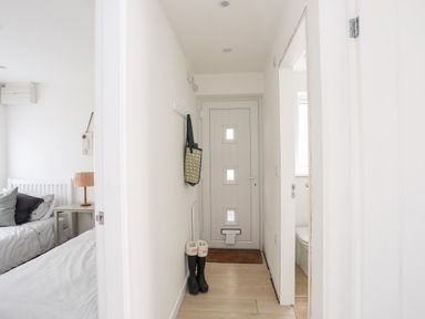 A narrow hallway with a white front door a pair of black boots a hanging bag visible bedrooms and bathroom at 31 Sisial Y Mor in Rhosneigr